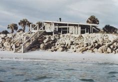 a house sitting on top of a pile of rocks next to the ocean with stairs leading up to it