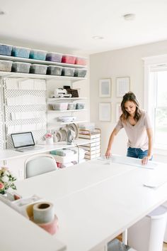 a woman standing in front of a white table with cups on it and shelves full of craft supplies