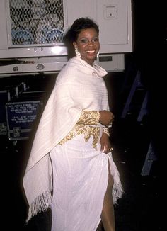 a woman in a white dress and gold jewelry standing next to a microwave with her hands on her hips