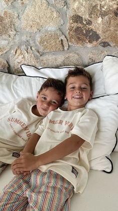 two young boys laying on top of a white bed next to each other in front of a stone wall