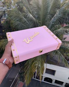 a person holding up a pink suitcase with gold lettering on it and palm trees in the background