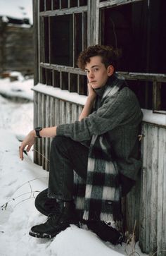 a young man sitting on the side of a building in the snow with his hand under his chin