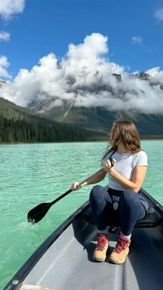 a woman sitting in a canoe on the water with mountains in the background and clouds in the sky