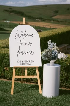 a welcome to our forever sign next to a white vase with flowers in it on the grass