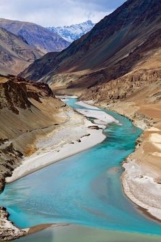 a river flowing through a mountain valley surrounded by snow capped mountains and blue water in the foreground