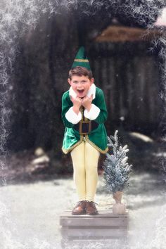 a young boy dressed in green and gold standing next to a small christmas tree