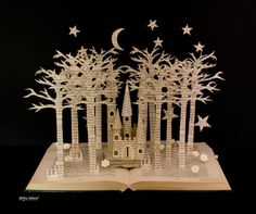 an open book with paper cut out of trees and stars on it, in front of a black background