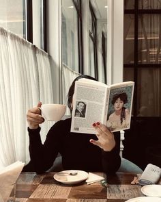 a woman sitting at a table reading a book and holding a coffee cup in front of her face