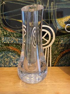 a clear glass vase sitting on top of a wooden table next to a wall painting