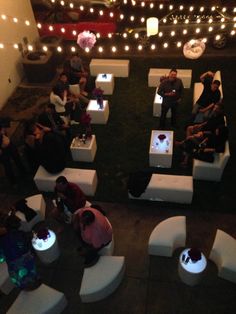 several people sitting on white couches in an open area with lights strung around them