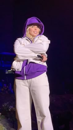 a woman in purple jacket and white pants standing on stage with her arms folded out