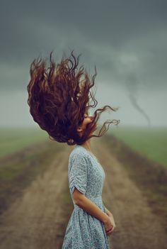 a woman standing in the middle of a dirt road with her hair blowing in the wind