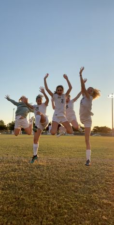 four girls jumping in the air with their hands up on a soccer field at sunset