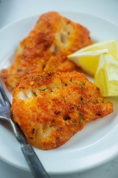 two pieces of fried fish on a white plate with lemon wedges and a fork