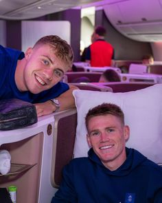 two men sitting in the aisle of an airplane, one is smiling at the camera