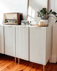 a white cabinet sitting on top of a wooden floor next to a mirror and potted plant