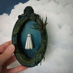 a hand holding a miniature white ghost in a frame with trees and clouds around it