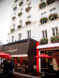 an outdoor cafe with red umbrellas and flowers on the windows, in front of a white building