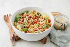 a white bowl filled with salad next to a wooden spoon and jar of dressing on a table