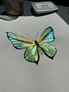 a green and yellow butterfly sitting on top of a white surface next to a cup