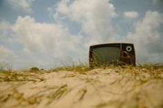 an old tv sitting in the sand with grass growing around it's edges and eyes