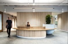 a woman sitting at a desk in an office with wooden walls and flooring, while a man walks by