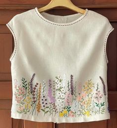 a white top with flowers embroidered on it hanging from a wooden hanger in front of a door