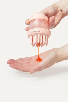 two hands holding a pink hair brush with orange liquid coming out of it, while the other hand is drizzling an orange substance