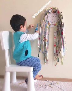 a little boy that is sitting on a chair with some scissors in his hand and paper clips hanging from the wall behind him