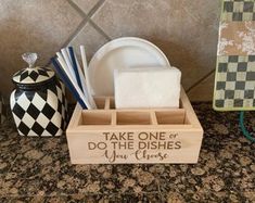 a wooden box with dishes and napkins in it on a kitchen counter next to a black and white checkered dish rack