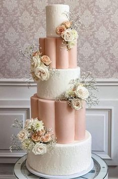 a three tiered wedding cake with pink and white flowers on the top, sitting on a table in front of a wall