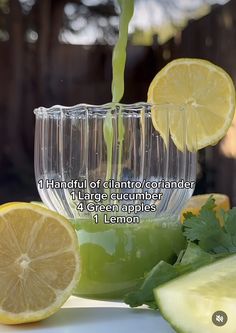 a glass filled with green liquid next to sliced lemons and cucumber slices