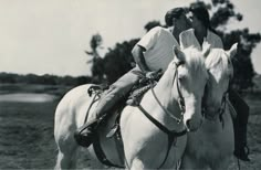 two men riding on the backs of white horses in a grassy field with trees behind them