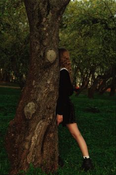 a woman standing next to a tree in the grass with her hand on it's trunk