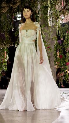 a woman is walking down the runway wearing a white gown and veil with flowers hanging from it