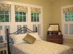 a bed sitting in a bedroom next to two windows with shutters on each side