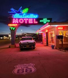 an old car parked in front of a motel at night with neon signs on the building