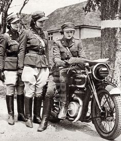 three men in uniforms are standing next to a parked motorcycle and another man is sitting on the bike