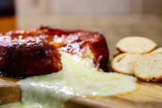 a wooden cutting board topped with cheese and meat covered in sauce next to crackers