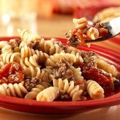 a red bowl filled with pasta and meat on top of a wooden table next to a fork