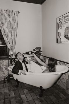 a man and woman laying in a bathtub drinking from wine bottles, with an advertisement on the wall behind them