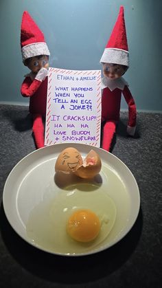 two elfs are sitting next to an egg on a plate with a sign in front of them