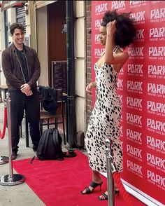 a woman in a dress standing next to a red wall with a man looking at her