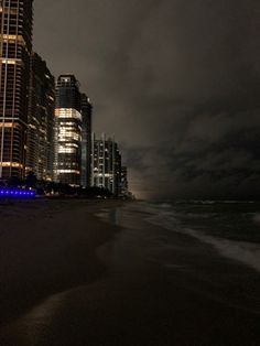 the city is lit up at night along the beach with dark clouds in the sky