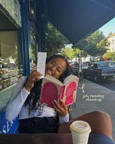 a woman sitting at a table reading a book and holding up a piece of paper