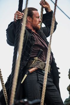 a man with a beard and leather jacket on top of a boat holding onto ropes