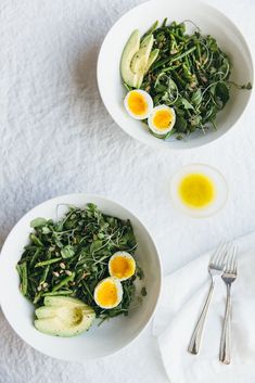 two bowls filled with greens and eggs on top of a white cloth next to silverware