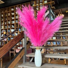 pink feathers are in a white vase on a wooden table next to bookshelves