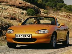 an orange sports car driving down a road next to a rocky hillside and tree covered hill