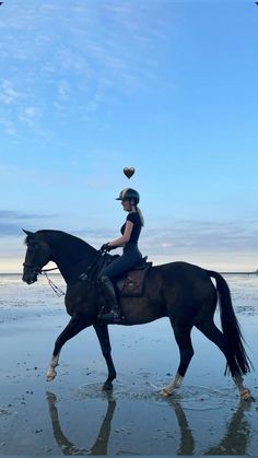 a woman riding on the back of a brown horse across a wet sandy beach under a blue sky